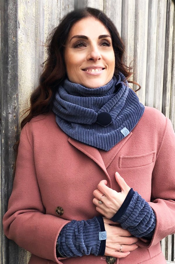 Frau trägt ein blaues Cord-Set aus Dreieckstuch mit Knopf und passenden Handstulpen, kombiniert mit einem rosafarbenen Mantel – gemütlich und stilvoll.  fair Wear  